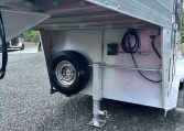 Underside view of a silver aluminum trailer with a spare tire, a deployed support jack, and hoses in a storage bay on a gravel lot.