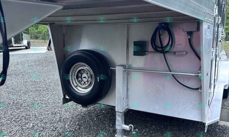 Underside view of a silver aluminum trailer with a spare tire, a deployed support jack, and hoses in a storage bay on a gravel lot.