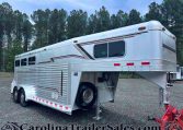 Silver aluminum gooseneck horse trailer with three axles parked on gravel, decorative stripes along the side, two windows, and a front hitch; trees in the background.