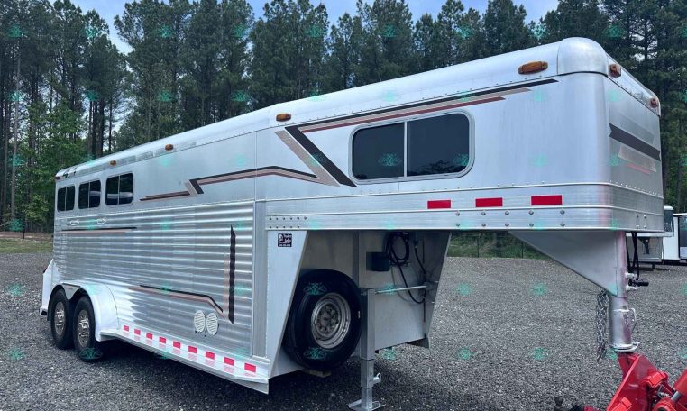 Silver aluminum gooseneck horse trailer with three axles parked on gravel, decorative stripes along the side, two windows, and a front hitch; trees in the background.