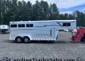 Long silver travel trailer on a gravel lot with tall pine trees in the background.