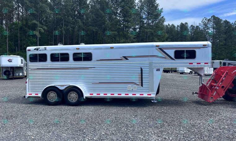 Long silver travel trailer on a gravel lot with tall pine trees in the background.