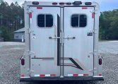Rear view of a silver double-door trailer with small windows, multiple latches and red reflectors, parked on a gravel lot with trees in the background.