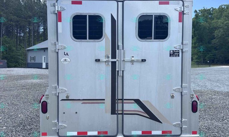 Rear view of a silver double-door trailer with small windows, multiple latches and red reflectors, parked on a gravel lot with trees in the background.