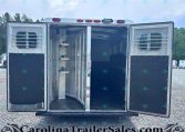 Rear of a silver horse trailer with both doors open, showing the interior stalls and divider bars, parked on a gravel lot.