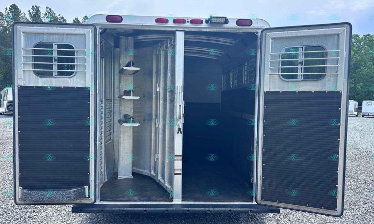 Rear of a silver horse trailer with both doors open, showing the interior stalls and divider bars, parked on a gravel lot.