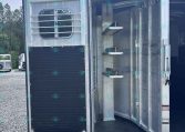 Rear of an aluminum horse trailer with the door open, showing interior with metal steps and a curved wall, parked on a gravel lot with other trailers in the background.