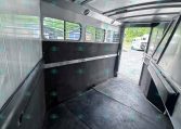 Interior of an empty aluminum horse trailer with windows and a rubber-lined divider, seen toward the open rear door.”