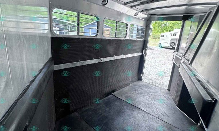 Interior of an empty aluminum horse trailer with windows and a rubber-lined divider, seen toward the open rear door.”