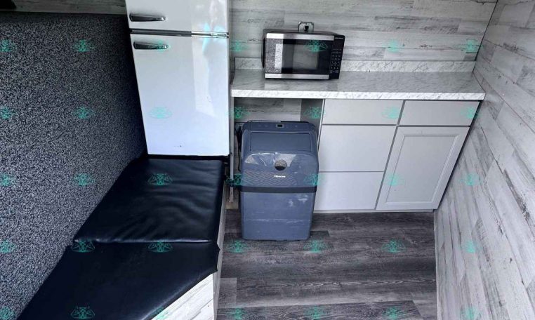 Trailer kitchen: white Galanz refrigerator with top freezer, microwave on a marble countertop, gray cabinets, and a trash can nearby.
