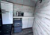 Compact kitchenette in a trailer: white retro refrigerator, gray upper cabinets, microwave on marble countertop, wood-paneled walls, and black hooks on the right wall