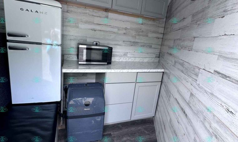 Compact kitchenette in a trailer: white retro refrigerator, gray upper cabinets, microwave on marble countertop, wood-paneled walls, and black hooks on the right wall