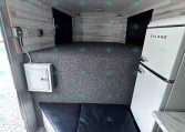 Interior of a small trailer living area with a white Galanz fridge on the right, black window curtains, gray speckled floor, and a black cushioned bench/sleep area. The walls and ceiling have light gray wood paneling and a round ceiling light.
