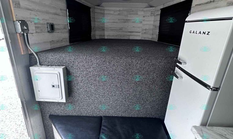 Interior of a small trailer living area with a white Galanz fridge on the right, black window curtains, gray speckled floor, and a black cushioned bench/sleep area. The walls and ceiling have light gray wood paneling and a round ceiling light.