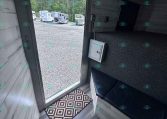 Interior of a small trailer with gray wood walls and seating; door open to a gravel lot with horse trailers outside.