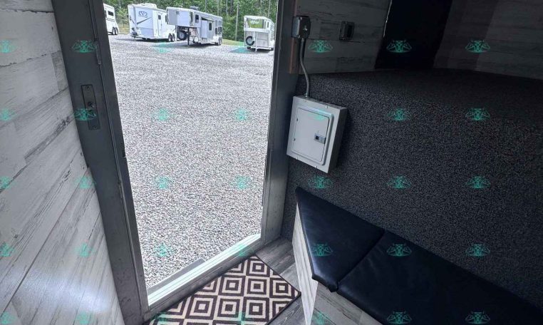 Interior of a small trailer with gray wood walls and seating; door open to a gravel lot with horse trailers outside.