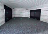 Interior of an empty trailer with a white ribbed ceiling, light gray wood-paneled walls, and two closed black roll-up doors at the ends.