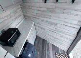 Compact trailer kitchen with white cabinets, a marble countertop, and a black microwave; gray wood-paneled wall features three black coat hooks and a diamond-pattern rug at the doorway