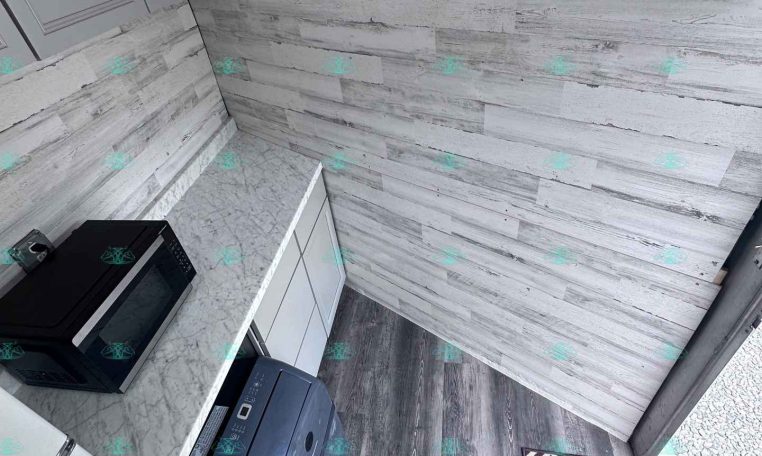 Compact trailer kitchen with white cabinets, a marble countertop, and a black microwave; gray wood-paneled wall features three black coat hooks and a diamond-pattern rug at the doorway