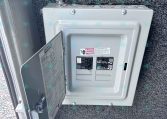 Open electrical breaker panel mounted on a textured exterior wall, with a conduit entering from the top and breakers visible inside the panel.