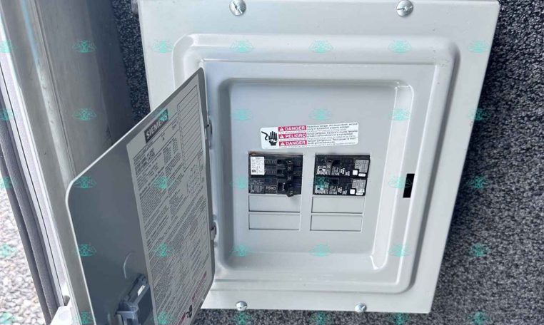Open electrical breaker panel mounted on a textured exterior wall, with a conduit entering from the top and breakers visible inside the panel.