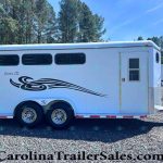 White horse trailer with three small side windows, decorative black swoosh graphics, and a rear door, parked on gravel with a forested backdrop; labeled Classic III.