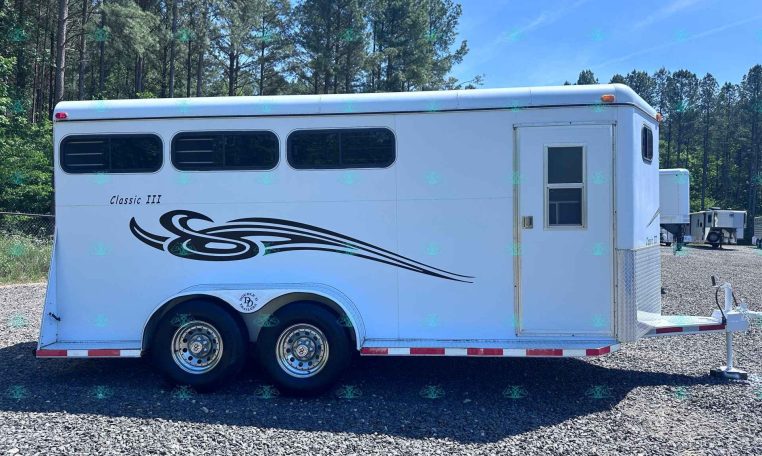 White horse trailer with three small side windows, decorative black swoosh graphics, and a rear door, parked on gravel with a forested backdrop; labeled Classic III.