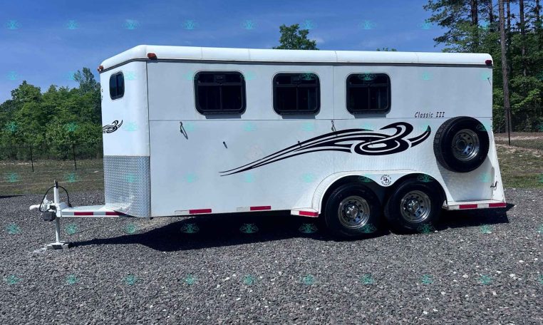 White enclosed horse trailer with three axles and chrome wheels on a gravel lot, trees in the background.