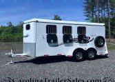 White horse trailer with three barred windows, spare tire on the side, and hitch on a gravel lot with trees in the background.