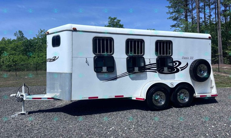 White horse trailer with three barred windows, spare tire on the side, and hitch on a gravel lot with trees in the background.