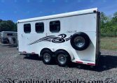 White horse trailer with black decorative swoosh, two small windows, and a spare tire mounted on the side in a gravel lot with trees in the background