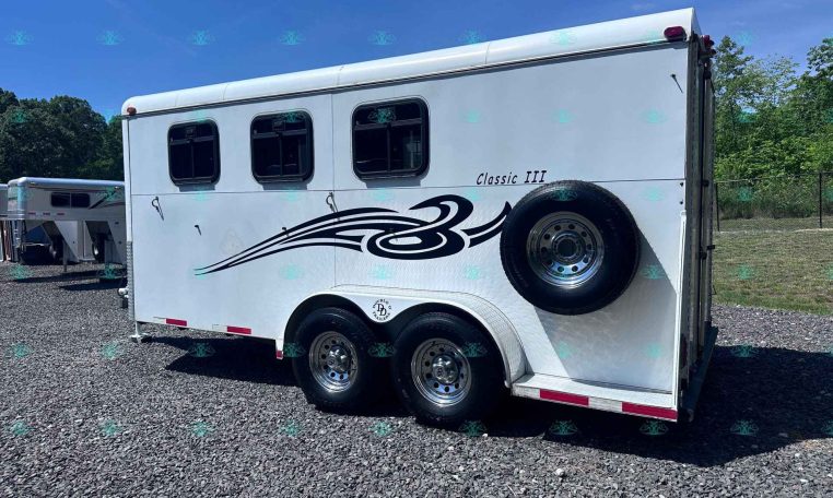 White horse trailer with black decorative swoosh, two small windows, and a spare tire mounted on the side in a gravel lot with trees in the background