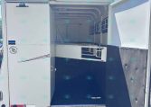 Open cargo trailer with rear doors swung open, exposing the white interior and blue floor inside a sunny lot with gravel outside.