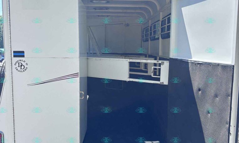 Open cargo trailer with rear doors swung open, exposing the white interior and blue floor inside a sunny lot with gravel outside.