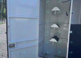 Open white cargo trailer with the door swung open, exposing an empty interior with built-in angled shelves and hooks against a bright outdoor backdrop.