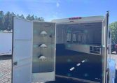 Open horse trailer with interior stalls and divider, parked on a gravel lot with trees in the background.