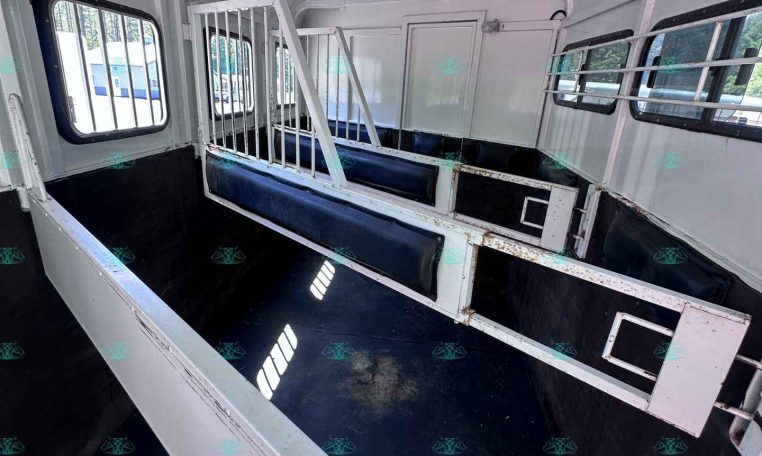 Interior of a white livestock trailer with metal stalls, blue padded benches, and barred side windows.