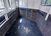 Interior of an empty horse trailer: white metal walls, black rubber flooring, padded side benches along the walls, barred windows, and a center divider.