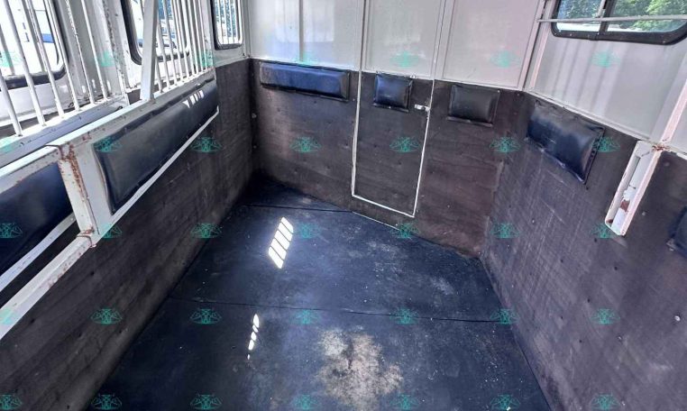 Interior of an empty horse trailer: white metal walls, black rubber flooring, padded side benches along the walls, barred windows, and a center divider.