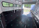 Interior of a horse trailer with black padded walls, rubber floor, and barred windows; rear ramp open to outside showing a trailer yard.