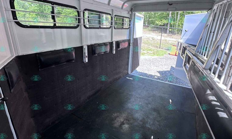 Interior of a horse trailer with black padded walls, rubber floor, and barred windows; rear ramp open to outside showing a trailer yard.