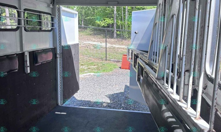 Interior of an empty horse trailer with stall dividers and rubber floor, open rear door to outdoors.