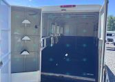 Interior of an empty horse trailer with partitioned stalls and saddle racks on the left wall; rear doors open and bright blue sky outside.