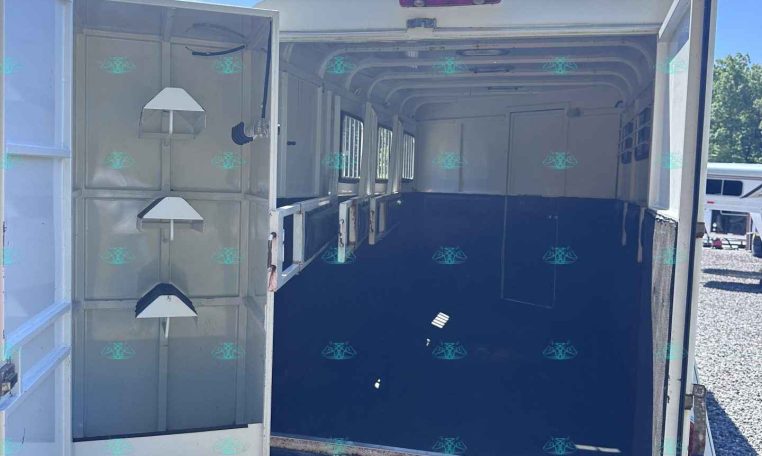 Interior of an empty horse trailer with partitioned stalls and saddle racks on the left wall; rear doors open and bright blue sky outside.