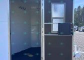 Open side door of a white trailer revealing an empty interior with wall hooks and a small cabinet; gravel lot and another trailer in background.