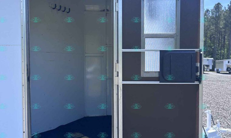 Open side door of a white trailer revealing an empty interior with wall hooks and a small cabinet; gravel lot and another trailer in background.
