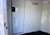 Interior of a small trailer or camper with a metal door, white walls, and a row of black hooks on the wall; a small window/vent is near the ceiling.