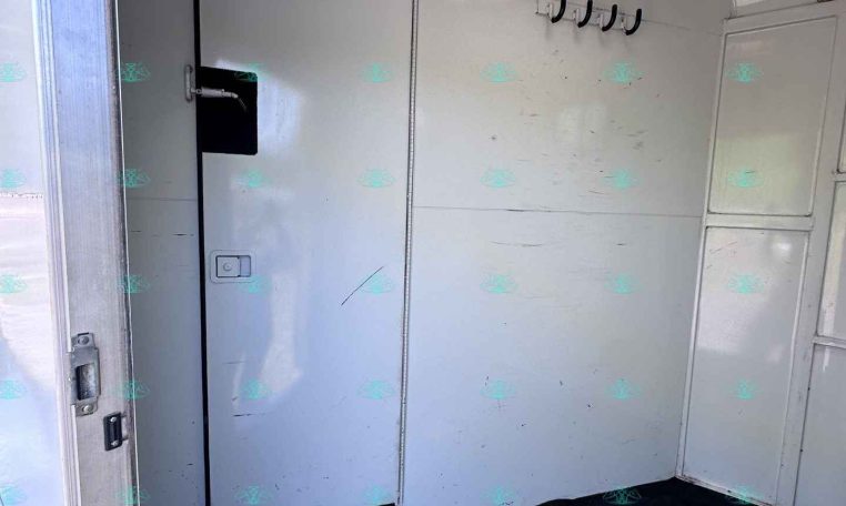 Interior of a small trailer or camper with a metal door, white walls, and a row of black hooks on the wall; a small window/vent is near the ceiling.