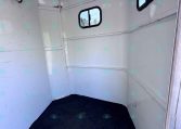 Interior of a white aluminum trailer with curved walls, a small window, and a dark carpeted floor with a few hooks on the left wall.