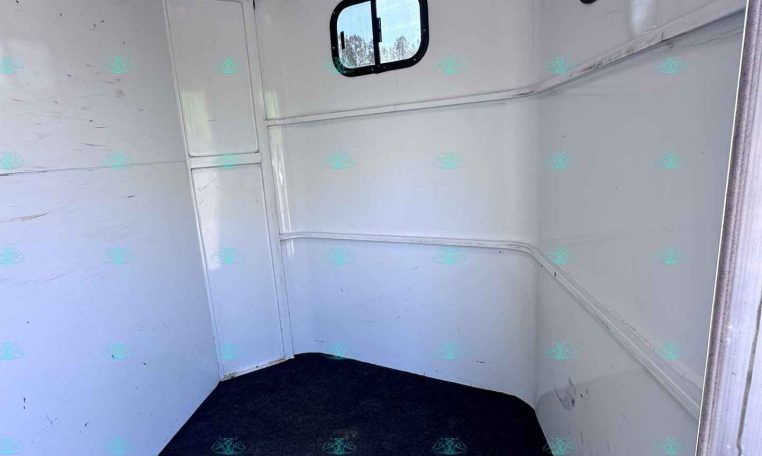 Interior of a white aluminum trailer with curved walls, a small window, and a dark carpeted floor with a few hooks on the left wall.